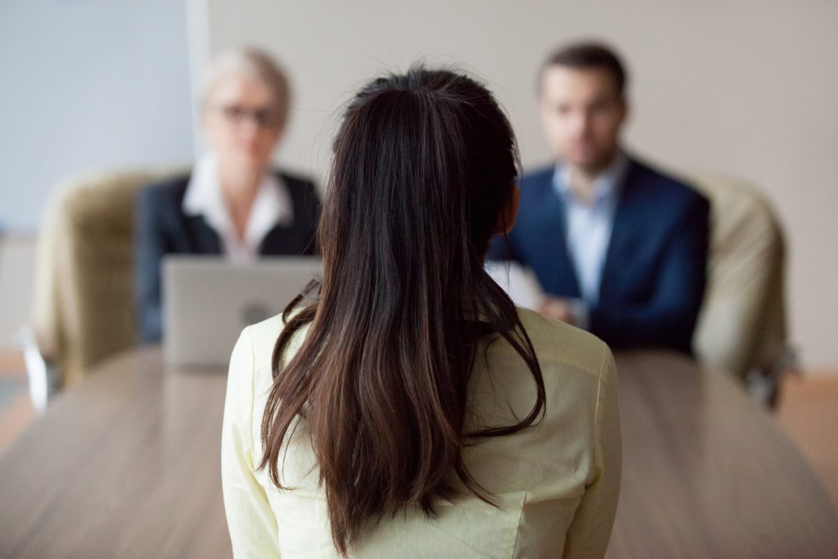 Businesswoman and businessman HR manager interviewing woman. Candidate female sitting her back to camera, focus on her, close up rear view, interviewers on background.